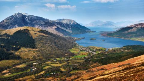 A hilltop view to Glencoe village and Loch Leven. Hills and mountains rise from the shores of the loch.