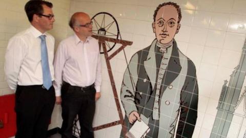 Two men looked at a tiled mural of a man in 19th century attire holding a book in front of a coal mine pitwheel and an church building