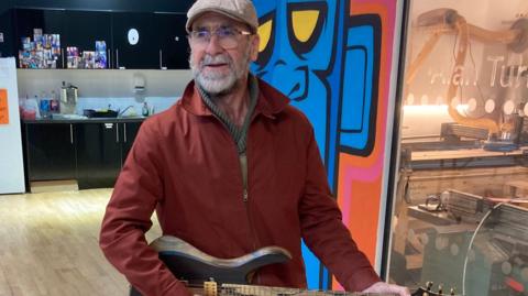 Eric Cantona holds a wooden guitar in GRIT artist studios