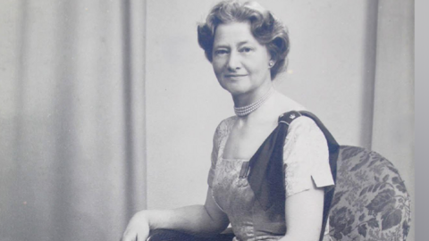 A black and white photo of a lady sat in an arm chair with a long dress on