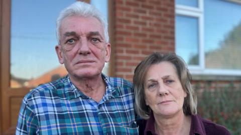A man, with short white hair, wearing a check shirt, standing beside a woman with shoulder length brown hair, wearing a purple top. They are stranding in front of a red brick house.