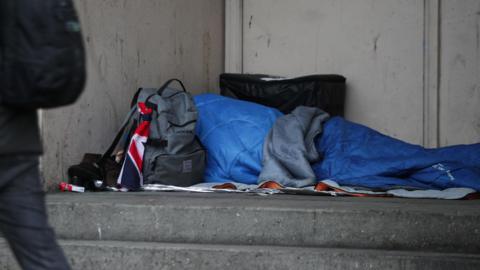 A homeless person sleeping rough in a doorway. They are in a blue sleeping bag with their belongings around them in other bags
