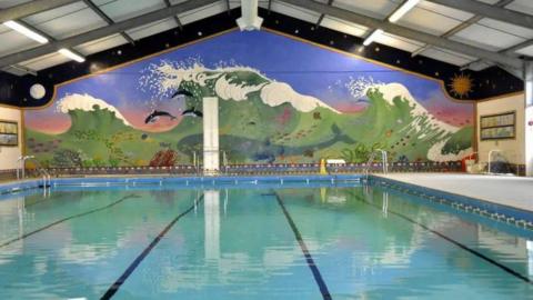 A swimming pool with a wall behind it that has a huge mural on it. The mural depicts a large green wave with a blue sky. There are dolphins leaping from the wave, a large whale is inside the wave. There are fish swimming through it and coral on the bottom.