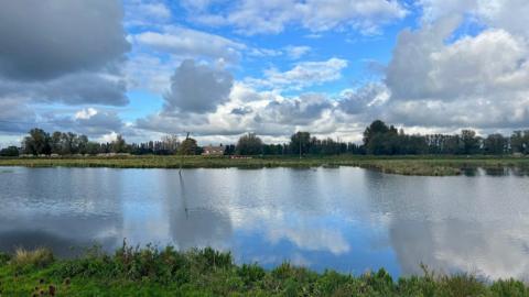 A wide expanse of river in a rural landscape under a partly cloudy sky