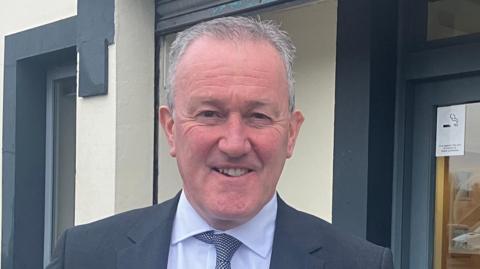 Minister Murphy wearing a dark grey suit with a grey and white tie, and a white shirt. He is standing in front of a white building with grey doors and windows and is smiling directly at the camera.