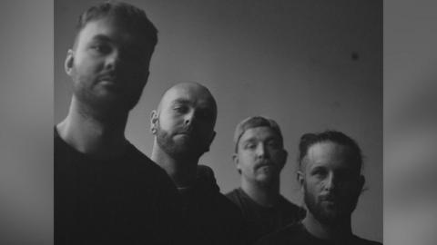 The four band members of Collapse The Sky face the camera, all of them unsmiling. They are photographed in a black and white and one of the them is wearing a cap. 