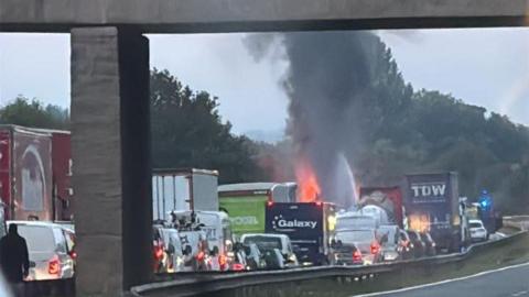 The fire seen from a distance with cars nearby queuing near it. A plume of smoke is seen coming from it. 