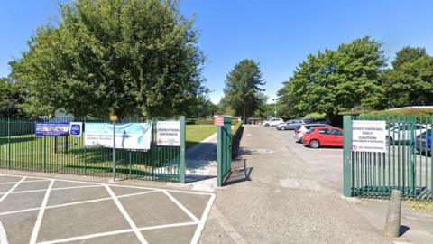 Primary school gates in green with a car park behind it and trees