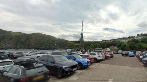 Cars parked in a row on the pier. behind them are railings and the quay. Grass covered cliffs are behind the water. The statue is in the centre. The statue depicts a pregnant woman holding aloft a sword while carrying the scales of justice and standing on a pile of law books