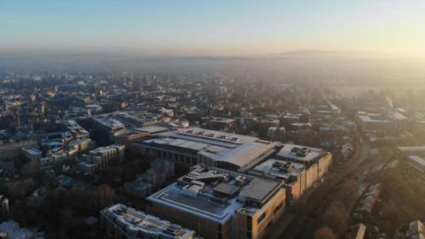 An aerial view of Oxford city on a winter morning. Spires are in the far distance, but it is mainly modern-looking buildings in the foreground.