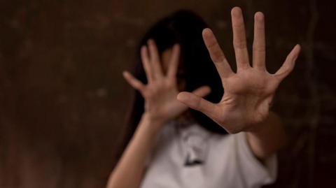 A woman standing with her hands out infront of her, spread out hands in a defensive position, she's looking away from the camera. She has long dark hair. 