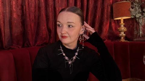 A woman wearing a black top, a necklace, and earings sits in a red booth, with red curtains in the background. She is looking to the left of the camera
