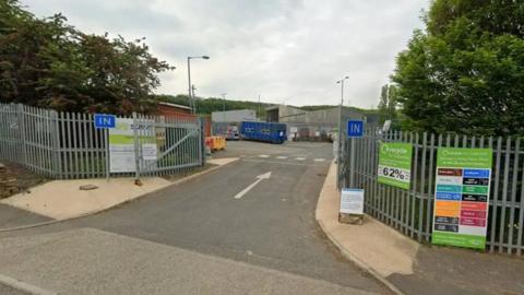 The entrance to a recycling facility with metal fencing, “IN” signs on both sides of the gate, and several information boards. A green sign promotes local recycling, while modern green‑and‑grey buildings stand behind the fenced area under a clear blue sky.