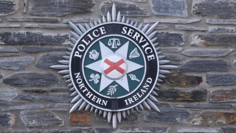 The badge of the Police Service of Northern Ireland is displayed against a stone wall