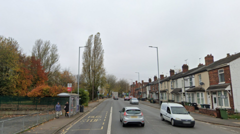 The road has two lanes and traffic can be seen using both sides. There are terraced houses on the right side of the image and a bus stop on the left with some people waiting there. 
