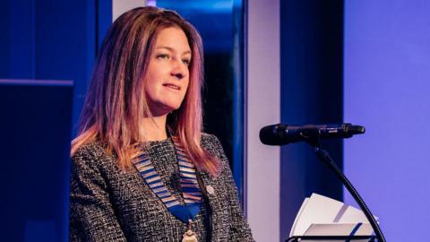Claire Watterson, who has long light brown hair and it wearing a black and white top, standing in front of a microphone at a podium. the panels in the background are purple and blue.