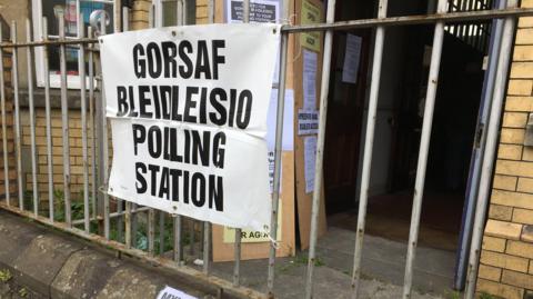 Bilingual sign saying "polling station/gorsaf pleidleisio" attached to railing outside a village hall. 