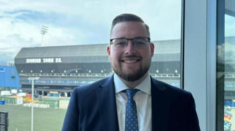 A man is dressed in a blue suit with a white shirt and a blue and white patterned tie. He is standing in the Suffolk County Council building with Ipswich Town Football Club in the background, through a window.