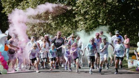 A group of men, women and children running together towards the camera. They are wearing sports clothing and googles, while pink and blue powder surrounds them.