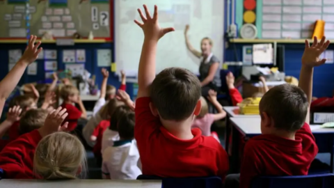 Pupils in a red uniform face a teacher with their backs to the camera and have their hands in the air as she speaks. There are about 15 boys and girl in the classroom with a teacher at the front, slightly blurred in the image.