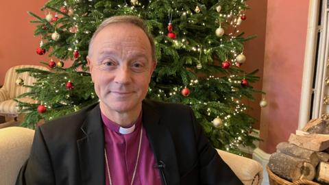 The Bishop of Exeter, Dr Mike Harrison, sat on a cream sofa. Behind him is a Christmas tree with red and gold baubles and strong lights. To the right of him is chopped up wood. He is wearing a purple shirt and black blazer. 