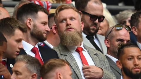 Preston Johnson in a grey suit, white shirt and red tie, sitting in the crowd at a Crawley Town football match