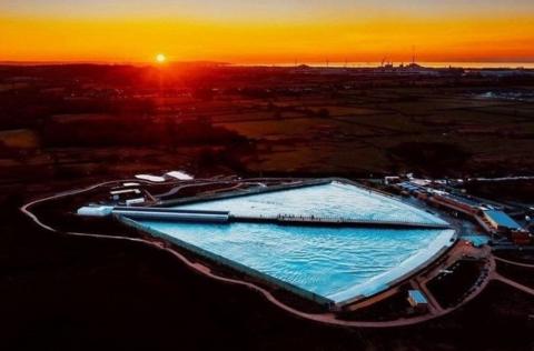 An aerial drone shot of The Wave, a man-made surfing lake on the outskirts of Bristol. It is taken at sunset with the sun just visible on the horizon. The water of the wave is light blue against the darkness around it