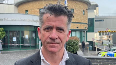 Michael Meredith, who is wearing a white shirt and grey suit jacket. He has short grey hair and is stood in front of the hospital building with a serious look on his face.