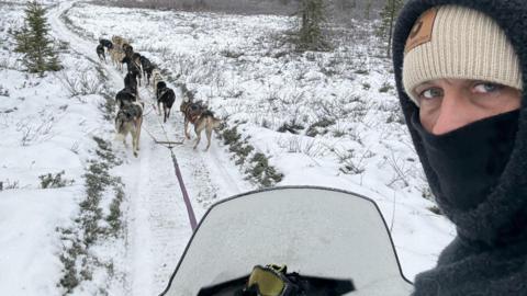 Danny Welch turning to look over his shoulder, with a grey fleece hood up covering a cream wool hat and a black balaclava pulled up to his nose obscuring most of his face. He is sitting on a sledge with a glass screen in a snowy landscape and ahead of him are a team of huskies pulling the sledge along a snowy track.