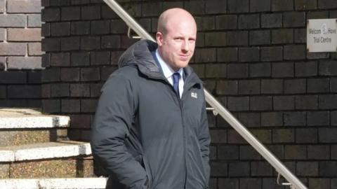 A bald man in a black coat and navy suit walking down stairs and looking at the camera.