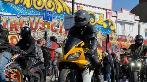 Several motorcyclists ride along Southend seafront past the arcades. All motorcyclists are wearing black helmets. The closest motorbike is mostly yellow in colour.