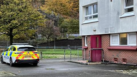 A police car parked outside Overtoun Court in Clydebank. The police car is white, with luminous yellow, red and blue detailing and 'Police' written on the side in blue text. The block of flats has exposed brick on the ground floor, with a red door and a window in the middle of two white grates. Above it, the building is light stone pebble dashed and has windows with white frames.