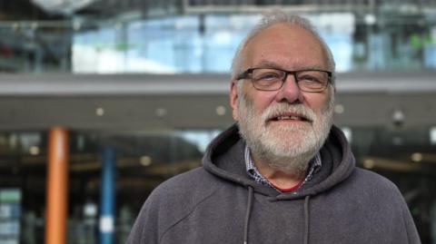 Steve Morphew is wearing a grey hoody with a yellow horizontal stripe across the chest. Beneath the top he is wearing a checked shirt.