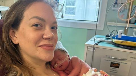 A mother in a hospital room with a newborn baby resting on her chest. She has long brown hair. The baby wears a knitted blue hat and nappy. There are various medical wires attached to the baby.