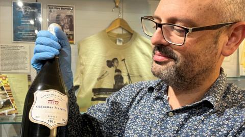 A man with short dark beard wearing a floral blue shirt holds a wine bottle. 