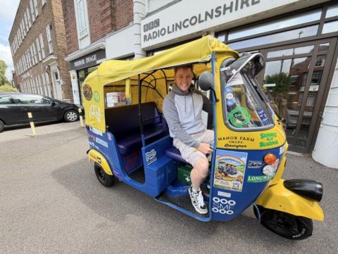 A man with a grey hoodie and shorts sits in a yellow and blue tuk-tuk.