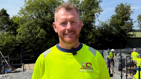 Chris Quinn, a man with ginger hair and beard, wearing a high-vis top on a building site.