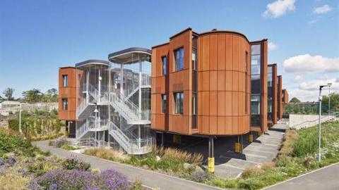 A modern brown building with exposed staircases surrounded by greenery and flowers.