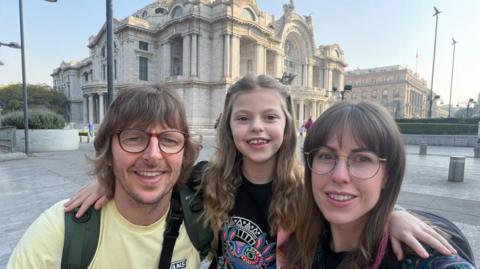 Craig, Summer and Alli Henry. They are posing for a selfie and smiling in front of a large marble building with highly decorated arches and columns. Crais has mid-length brown hair and glasses with a round red frame and blue eyes. He is wearing a pastel yellow t-shirt and a rucksack. Summer is in between her parents and has her arms around their shoulders. She has shoulder-length blond hair and brown eyes. She is wearing a black top with a fluorescent graphic on the front. Allu has long brown hair and a fringe. She wears round glasses with a metal frame and a multicoloured top.