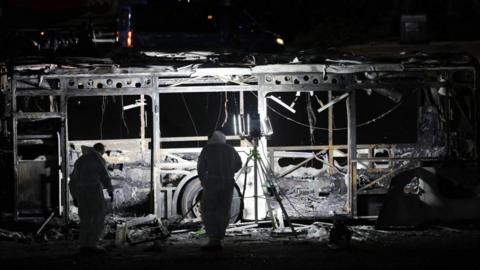 Bus burnt out with two people in white suits searching the wreckage using large light