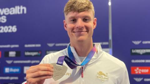A man holding a silver medal with a purple ribbon around his neck. He is wearing a white Team England tracksuit top