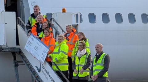 People in high-vis jackets stand on the stairs to a plane. Some of them are holding a large boarding pass. They are looking off to the left of the camera and smiling.