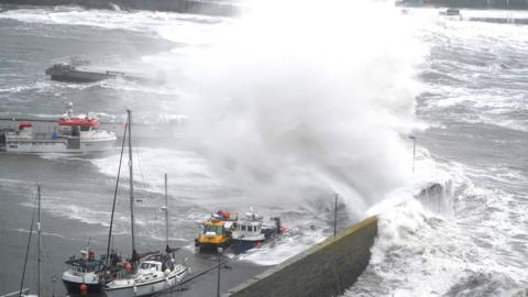 Large white waves crashing into a harbour over small boats that are tied up