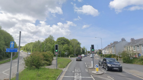 A google street view image of Barncoose Terrace. There is a road going up to the left and a road straight ahead. There is green grass and a pedestrian footpath in the middle of the two. There are cars on the road driving in both directions. There are traffic lights which are green. There are houses on the right. 