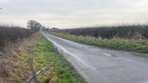 A single lane road stretches into the distance. It is flanked by overgrown grass verges and large hedges. The sky is grey and cloudy.