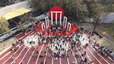  An aerial view shows crowds gathered around the Shaheed Minar, a monument with five rectangular structures, the one in the middle larger, on a red plinth and with a red disc behind it. People are gathered around the steps in front of it, with flooring that is reddish brown with circular mosaics.