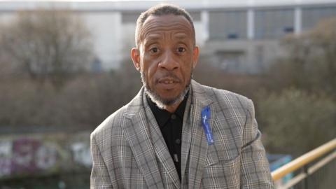 A black man looks directly into the camera with soft-focus workshop units in the background