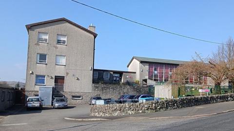 A side view of an old secondary school part of which is on three storeys with a long, glass-fronted annex attached. There are cars parked outside and a lovely blue sky above