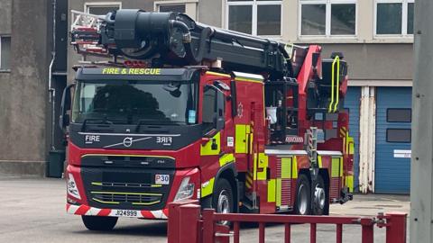 A red fire engine with yellow markings on the side and an aerial pump stored on the roof. It is parked at the fire station.