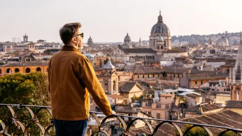 A man looks out from a balcony over rooftops and the dome of St Peter's Basilica in Rome